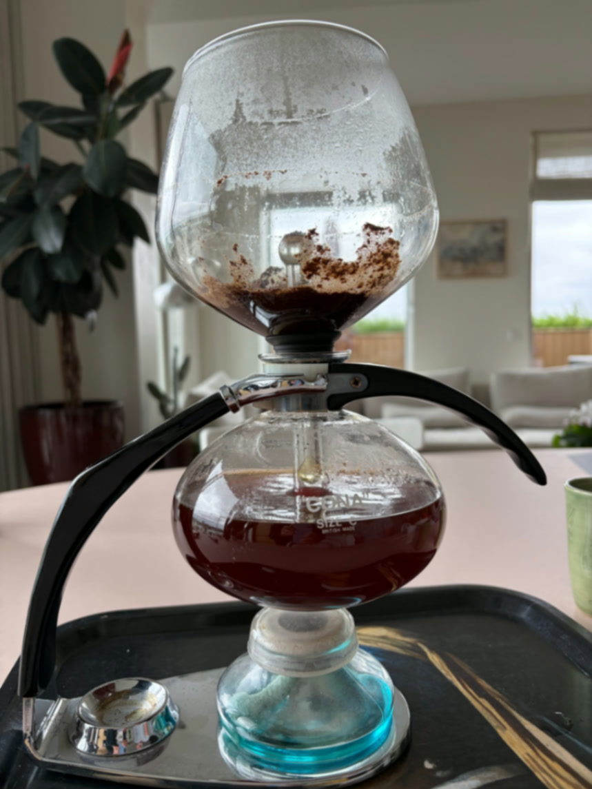A vintage Cona vacuum siphon brewer mid-extraction on a metal tray